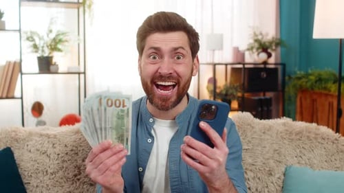 Excited man holding money and phone while sitting