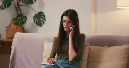 Woman Taking Notes on Couch While Talking on Phone