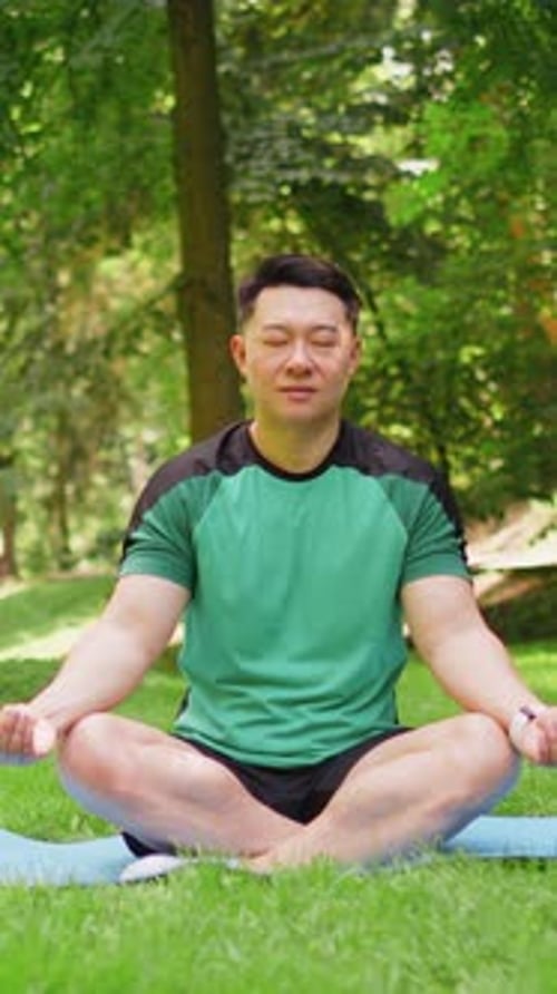Young Asian Man Sitting on Mat in Lotus Position Relaxing Practicing Yoga in Park on Summer Day