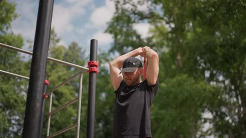 Outdoor Park Male Stretching Arms Overhead Cap Beard Black Shirt at Pullup Bar Under Leafy Trees