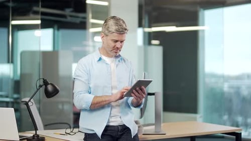 Mature business man uses a digital tablet standing in a modern office indoors. Gray-haired senior