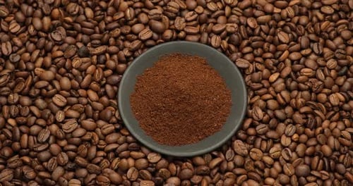 Keramische Untertasse voller gemahlener Kaffeebohnen auf rotierendem Hintergrund aus frisch gerösteten Kaffeebohnen