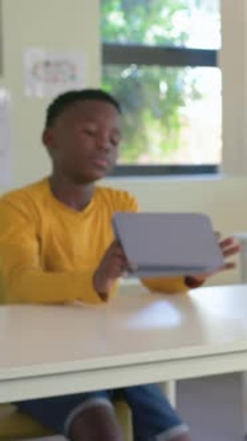 Vertical video: Raising head after pause yellow-shirt boy checking tablet at classroom cam panning