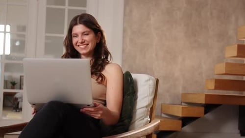 Eine langhaarige Brünette winkt mit der Hand vor der Webcam eines Laptops und fängt an, etwas zu sagen, jung