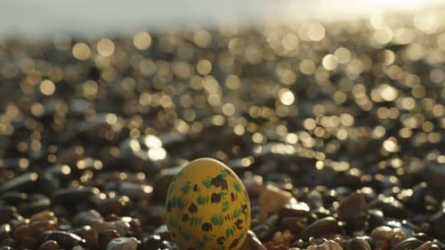 Ein bemaltes gelbes Ei zu Ostern, auf den Felsen am Meer. Der Fokus verlagert sich auf die Wellen und