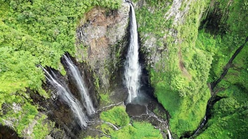 Aerial drone view of multiple narrow waterfalls flowing down steep green cliffs, surrounded by dense
