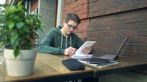 Happy Boy with Smartphone and Notepad Doing Homework by Table in Cafe Adolescent
