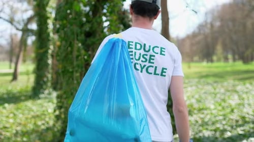 Man at plastic garbage collecting in a polluted park. Rubber gloves, carrying a bag with plastic bot