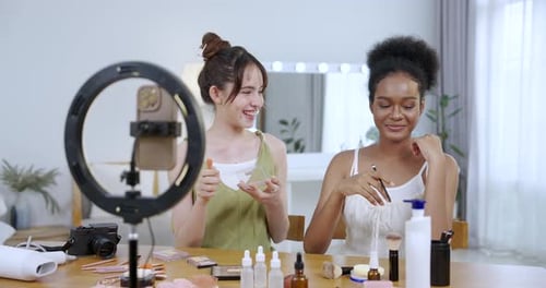 vloguera de belleza adolescente asiática sentada junto a una adolescente afroamericana sonriendo aplicándose cuidado de la piel