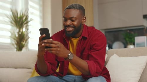 Close Up Happy Young African American Man Use Phone Smiling Read Good News Sit on Sofa in Home