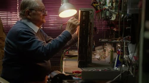 Elderly caucasian man fixes radio equipment in dark room, side view