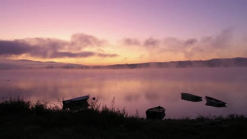 Dawn at a lake with amazing colours, anglers boats by a shore, a mist floating over a water surface.
