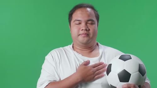 Asian Man With A Ball Putting Hand On His Heart To Respect While Cheering Soccer On Green Screen