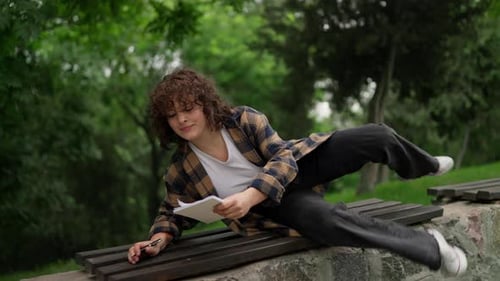 Confident Brunette Girl with Curly Hair Lies Down on a Bench While Writing Down Her Ideas and