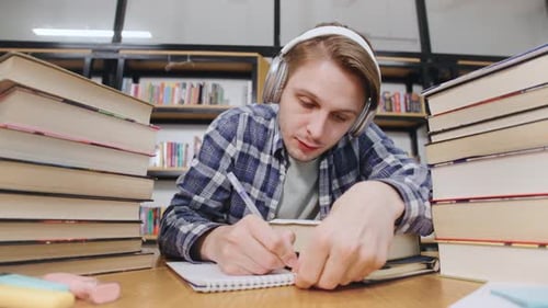 Student studiert in einer Bibliothek mit Bücherstapeln und Kopfhörern, während er an einem Holzblock Notizen macht