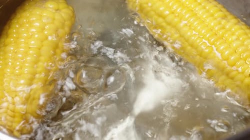Two corn cobs are boiling in bubbling water, captured from a top-down view. The slider moves
