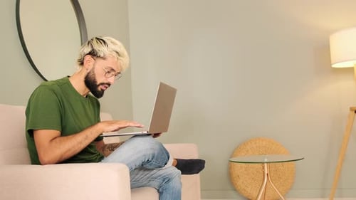 Young blond man with a beard works on a laptop while sitting in a cozy living room