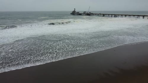 Drone footage of a pier in the ocean shore. Shot on a cloudy winter day. Lots of foam in waves. Dark