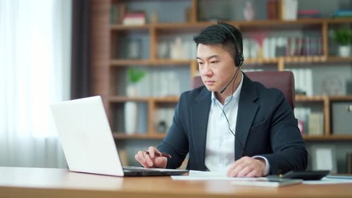 Asian employee studying online virtual tutor using laptop in office. distant worker wears headphones