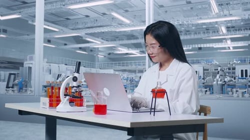 Asian Woman Scientist Typing On A Laptop While Working With Microscope And Test Tube In The Lab