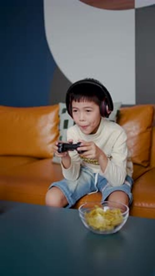 Junger asiatischer Junge genießt seine Freizeit, spielt mit Joystick auf einem Sofa und isst ein paar Snacks