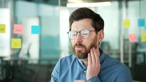 Upset businessman suffering from toothache sitting at workplace in business office. Bearded man