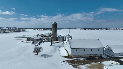 Snowy farm with silo, barns, and outbuildings, surrounded by open fields under a blue sky. Aerial or