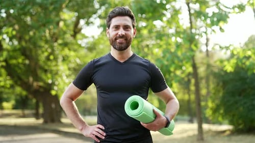 Porträt eines lächelnden bärtigen, athletischen Mannes, der mit einem selbstbewussten Lächeln draußen im Stadtpark steht