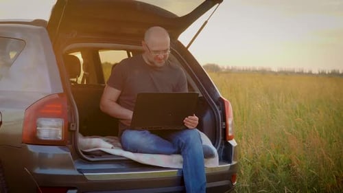 Empresário sorridente conversando com o cinegrafista fazendo uma conferência de negócios sentado no porta-malas do carro enquanto