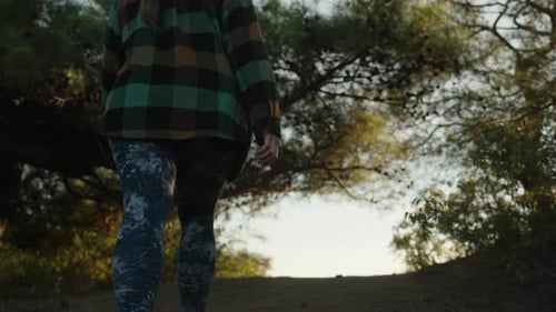 Eine junge Frau in Leggings und kariertem Hemd klettert einen Hang im Wald hinauf, um den Sonnenuntergang zu beobachten.