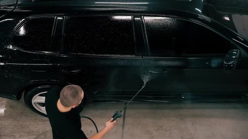 A male car wash employee applies car wash detergent to a black luxury car using spray gun in the car