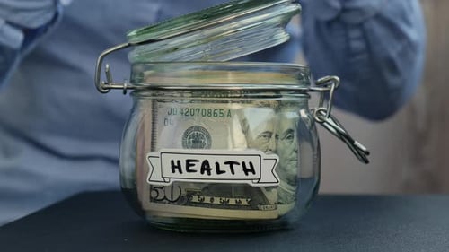 Female Hands Add Money to Saving Money In Glass Jar Filled with Dollars Banknotes HEALTH