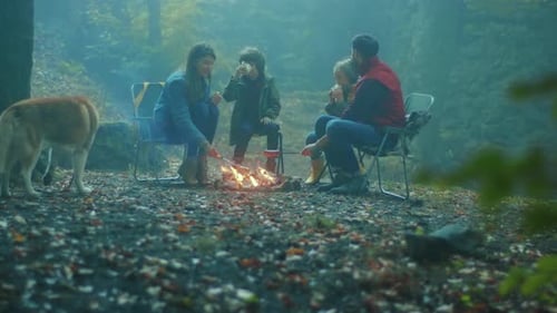 Family with Dog Sit Around a Campfire on Autumn Evening in the Woods Frying Sausages Over a Fire in