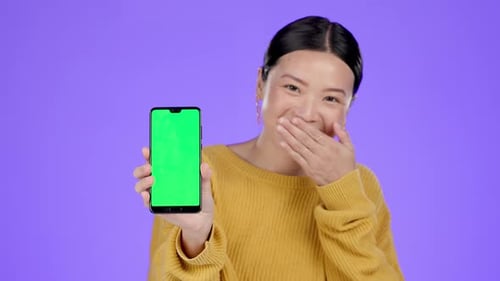 Asian woman, smartphone and green screen mockup in studio with hand on mouth for funny secret by