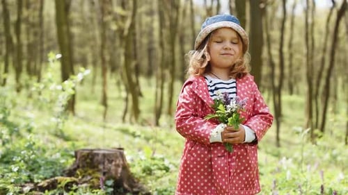 Cute little girl in blue hat standing in forest and holding flowers in hands