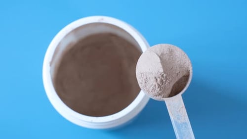 Hand takes out protein powder in scoop from jar on blue background top view.