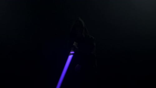 Young Man in Jedi Cosplay Costume with Lightsaber Battle on Black Background in Smoke and Rain Slow