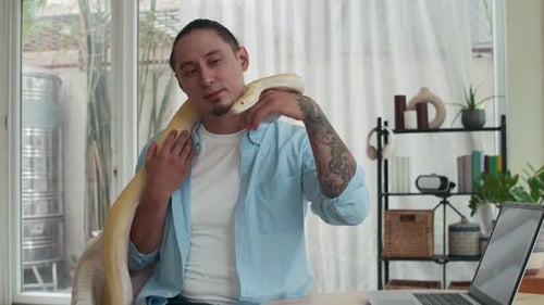 Homme assis avec un serpent albinos enroulant le cou pendant qu'il travaille à la maison
