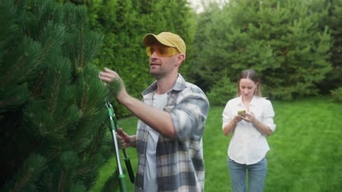 Un homme taille un arbre tandis qu'une femme utilise un smartphone à l'extérieur