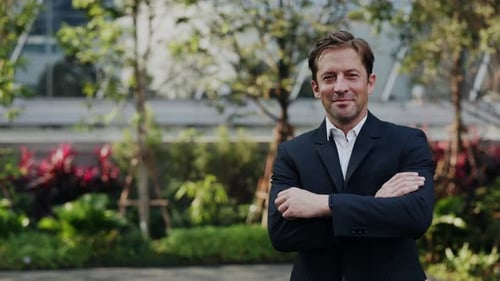 Portrait of Businessman Smiles and Crosses Arms in Blooming Urban Park