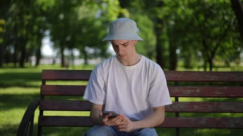 A Guy in a Sun Hat Sits on a Bench on a Sunny Summer Day in a City Park and Fussily Looks at the