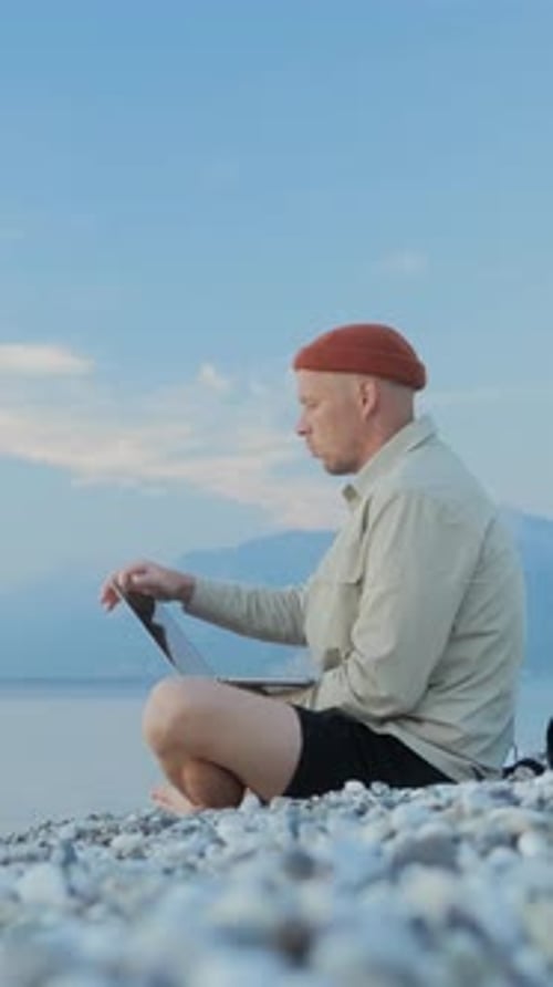 Weitwinkelaufnahme eines Mannes, der am Strand sitzt und bei Sonnenaufgang seinen Laptop öffnet