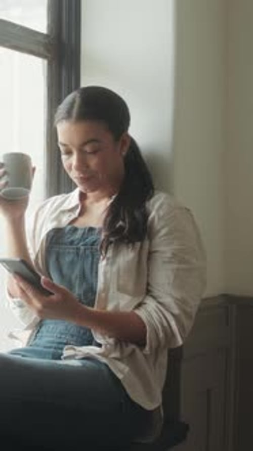 Woman Drinks Coffee and Uses Phone by Window