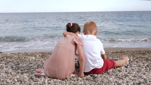 Kids Sit on the Beach By the Sea in an Embrace and Throw Pebbles Into the Water