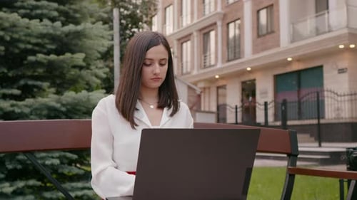 Woman sitting on bench at city street using laptop.