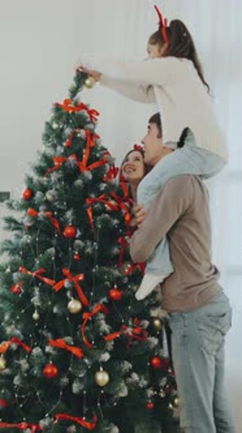 Família decora árvore de Natal com enfeites e fitas festivas em uma aconchegante sala de estar durante