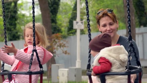 Kindheit, Spaß - junge Mama auf dem Spielplatz schiebt ihre Kinder auf die Schaukel