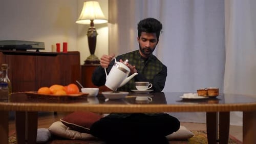 Plan large d'un jeune homme du Moyen-Orient assis sur des coussins à une table basse en train de verser du thé dans une tasse, vue de face