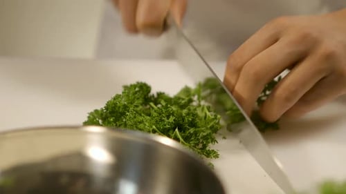 A chef's hand chops fresh parsley on a white cutting board with a sharp knife