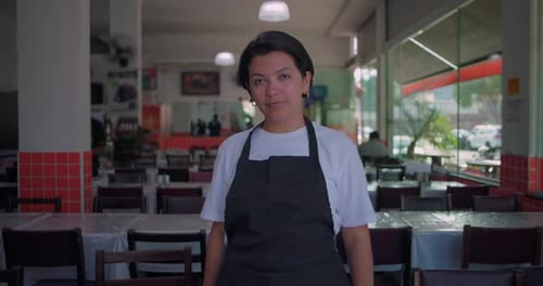 Latina waiter standing in restaurant dining area, looking directly at camera with calm and neutral